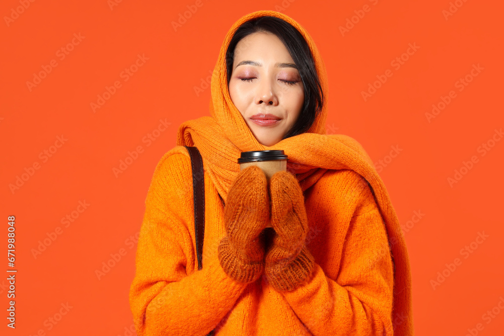 Young Asian woman in winter clothes with coffee cup on orange background