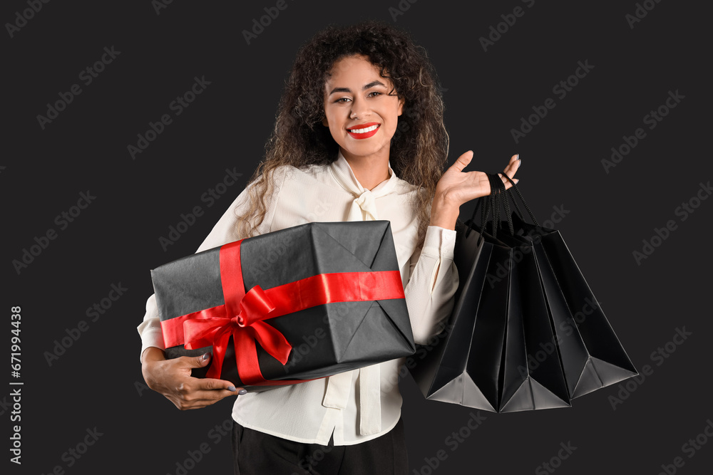 Beautiful African-American woman with shopping bags and gift box on dark background. Black Friday sale