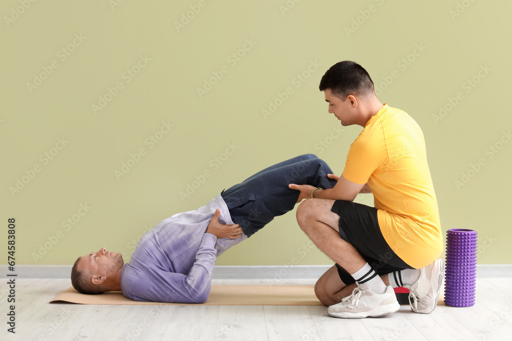 Mature man training on mat with rehabilitation therapist near green wall