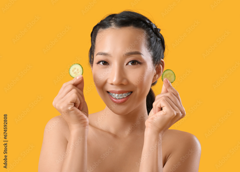 Young Asian woman with glass of cucumber water on yellow background, closeup