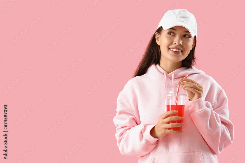 Beautiful young Asian woman in stylish hoodie with cup of lemonade on pink background