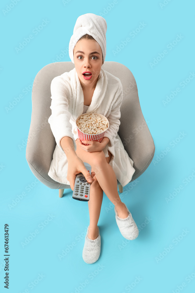 Emotional young woman in bathrobe with popcorn and TV remote control watching movie on blue background