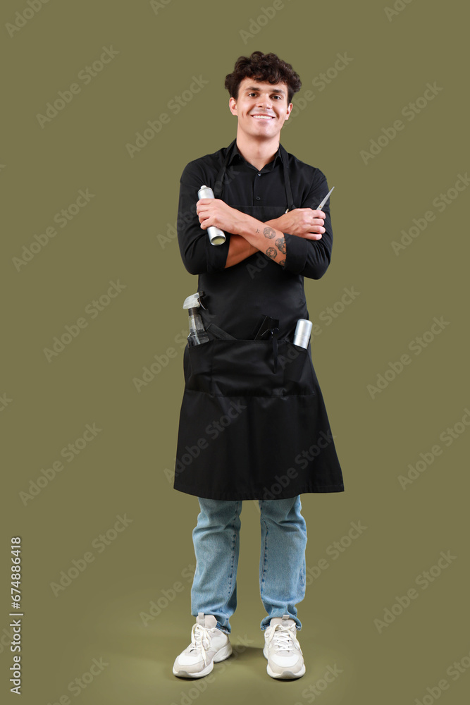 Handsome male hairdresser in black apron with scissors and hair spray on green background