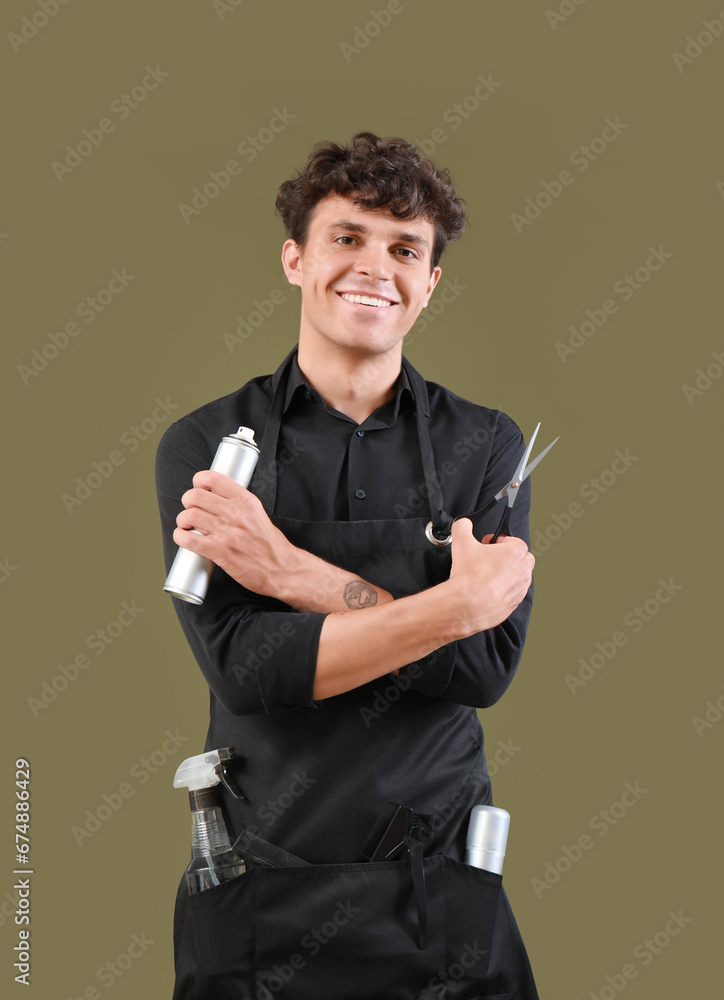 Handsome male hairdresser in black apron with scissors and hair spray on green background