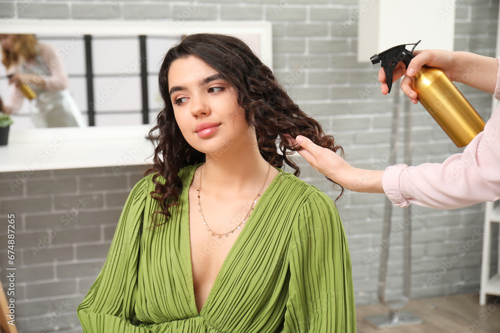 Professional hairdresser applying hair spray on young womans curly hair in beauty salon