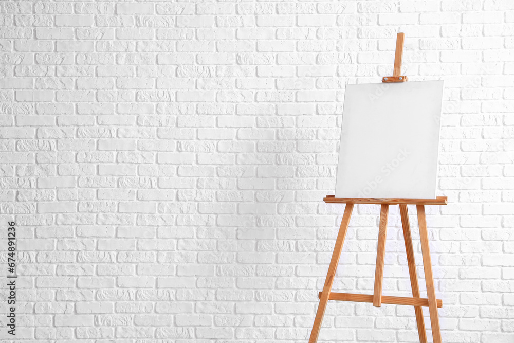 Wooden easel with blank canvas near white brick wall