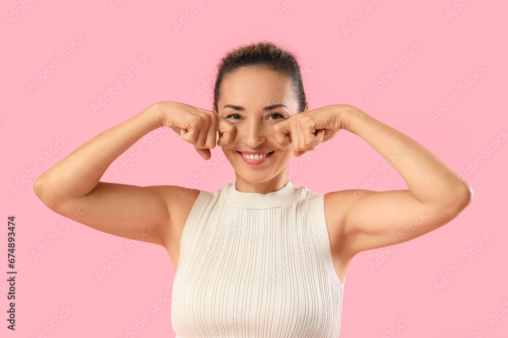 Beautiful mature woman giving herself face massage on pink background