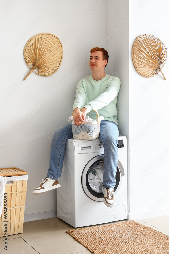 Young man with laundry basket sitting on washing machine at home