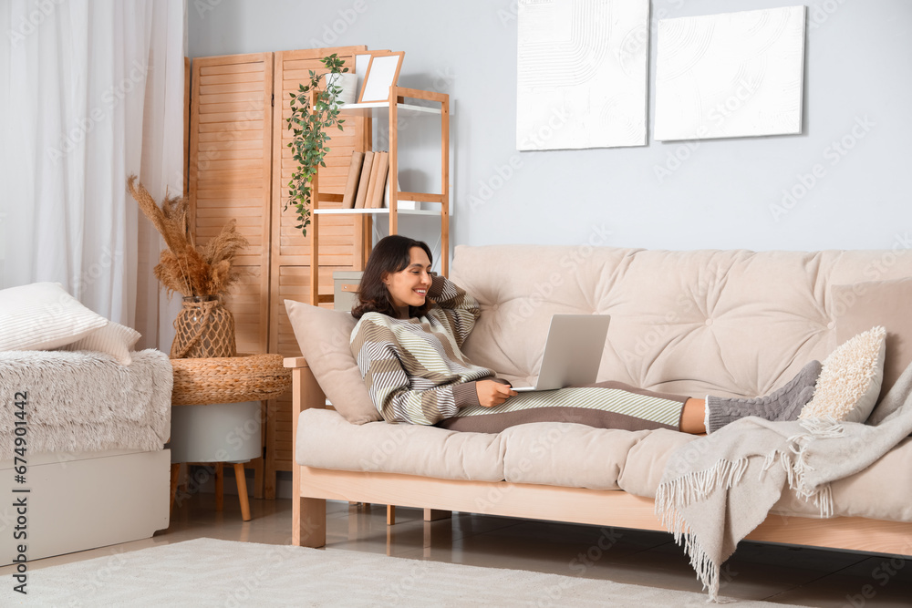 Young woman in warm sweater using laptop on sofa at home