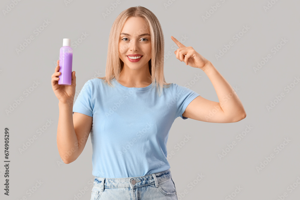 Young blonde woman with bottle of cosmetic product pointing at her hair on light background