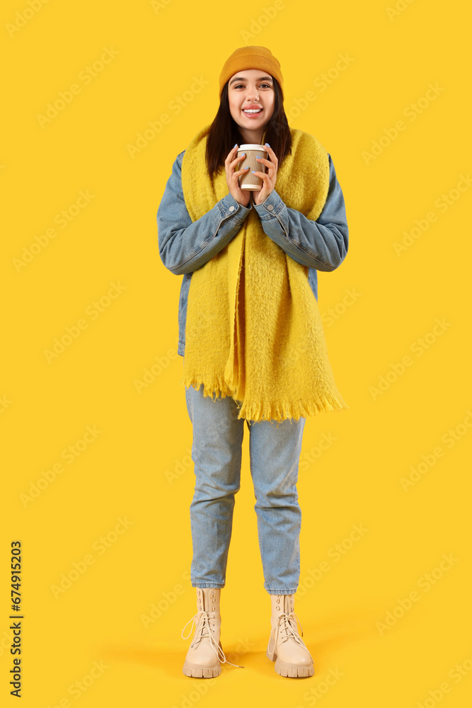 Young woman in winter clothes with coffee cup on yellow background