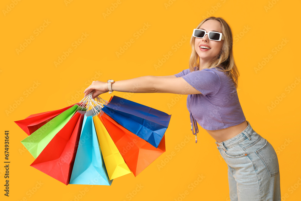 Young woman in sunglasses with shopping bags on yellow background