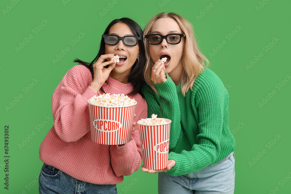 Beautiful young women in 3D glasses with buckets of popcorn on green background