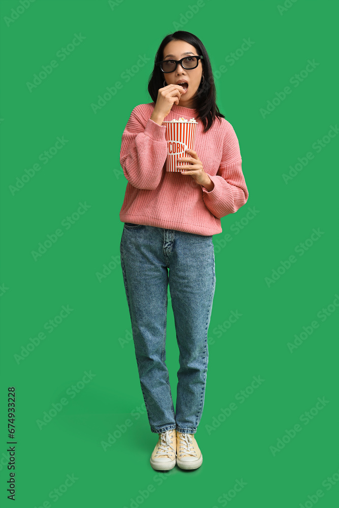 Young Asian woman in 3D glasses with bucket of popcorn on green background