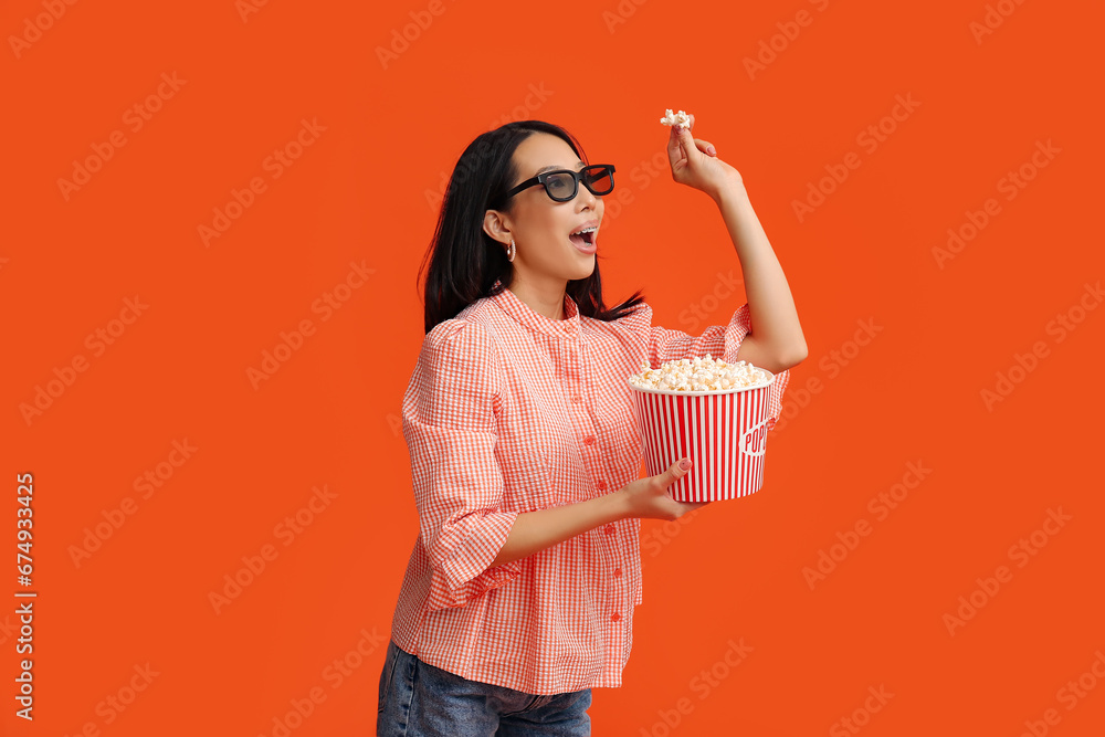 Young Asian woman in 3D glasses with bucket of popcorn on orange background