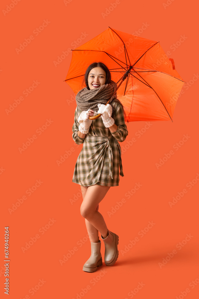 Beautiful young Asian woman in warm scarf with umbrella on orange background