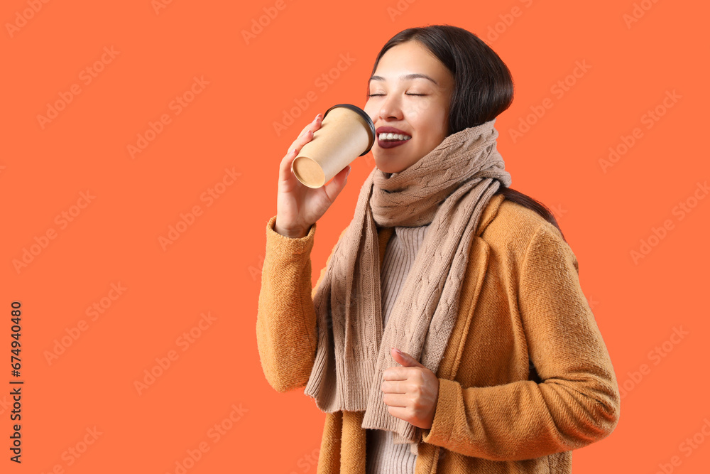 Beautiful young Asian woman in warm scarf with cup of coffee on orange background