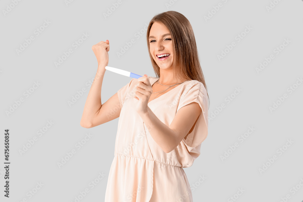 Happy beautiful young woman with pregnancy test on grey background