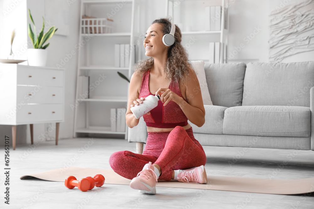 Smiling adult woman in sportswear with bottle of water sitting on yoga mat at home