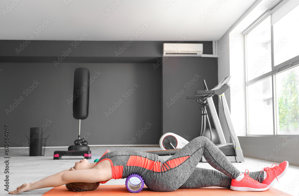 Beautiful young woman in sportswear doing exercises with foam roller at gym