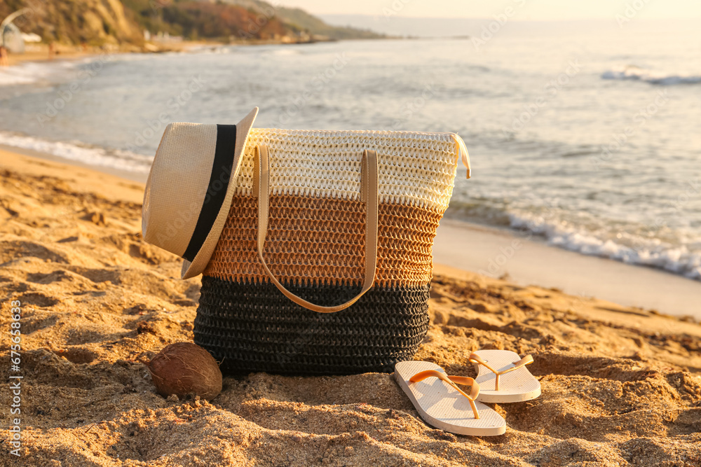 Stylish beach bag, coconut, hat and flip-flops on sand near sea