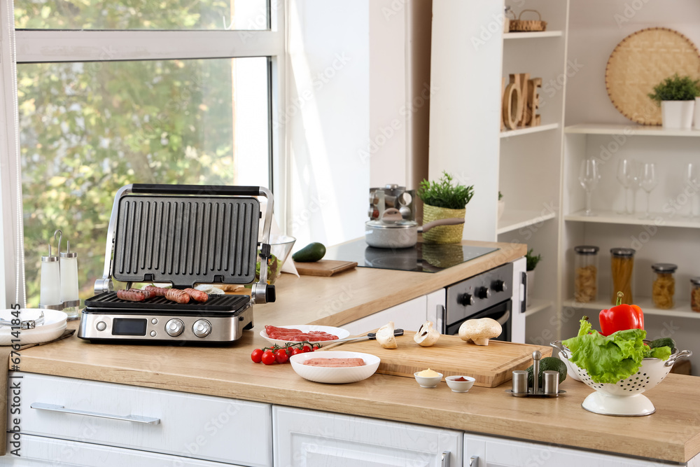 Modern electric grill with tasty sausages on wooden countertop in kitchen