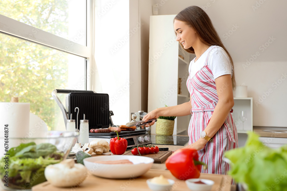 Beautiful young woman cooking delicious sausages on modern electric grill in kitchen