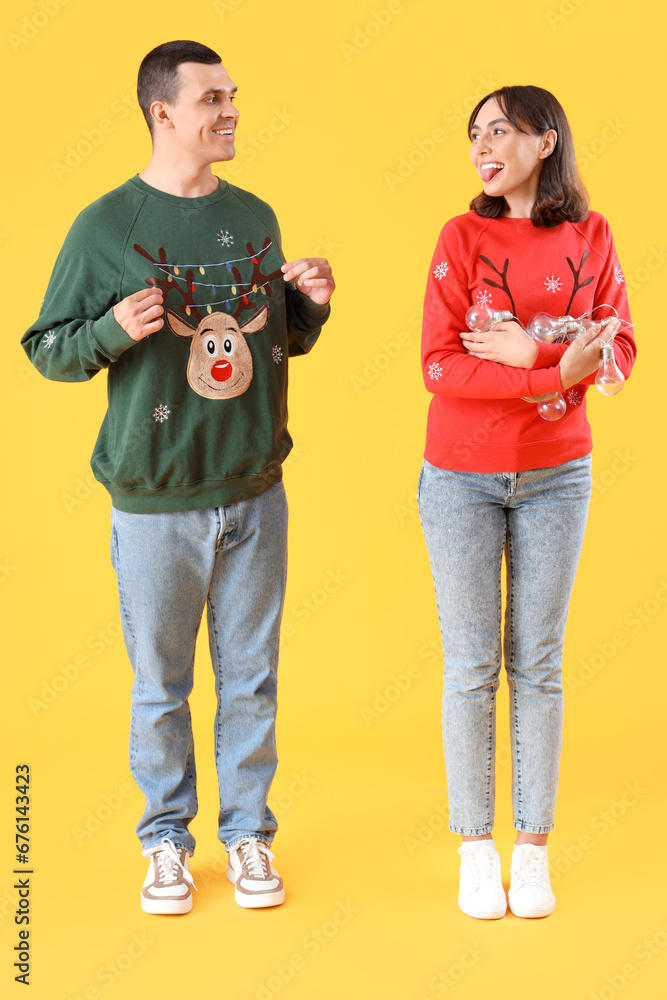 Young couple in Christmas clothes with bulb garland on yellow background