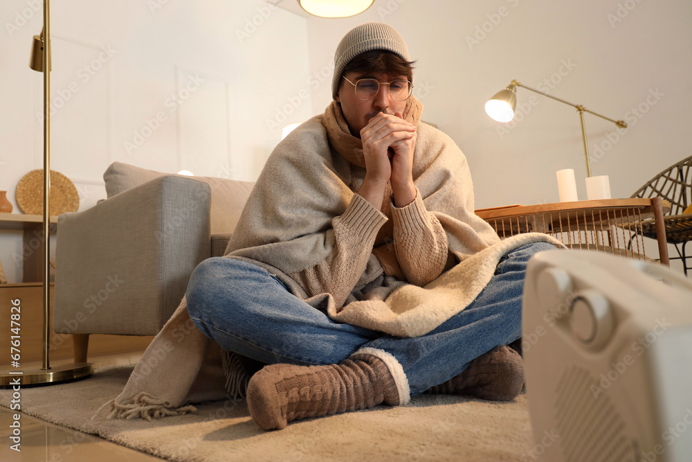 Young man warming near electric heater at home in evening