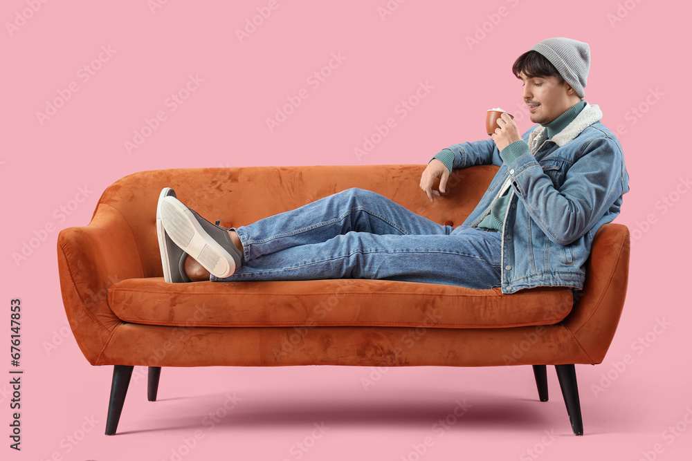 Young man in winter clothes with cup of cocoa lying on red sofa against pink background