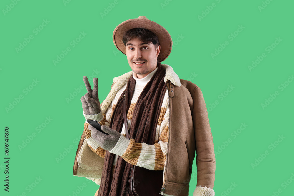 Young man in winter clothes with mobile phone showing victory gesture on green background