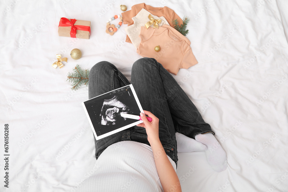 Pregnant woman with sonogram image, test, baby clothes and Christmas decorations on bed