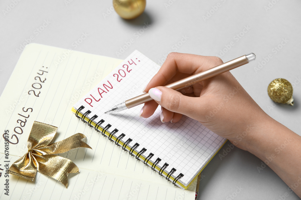 Woman writing PLAN 2024 in notebook and golden Christmas balls on grey background, closeup