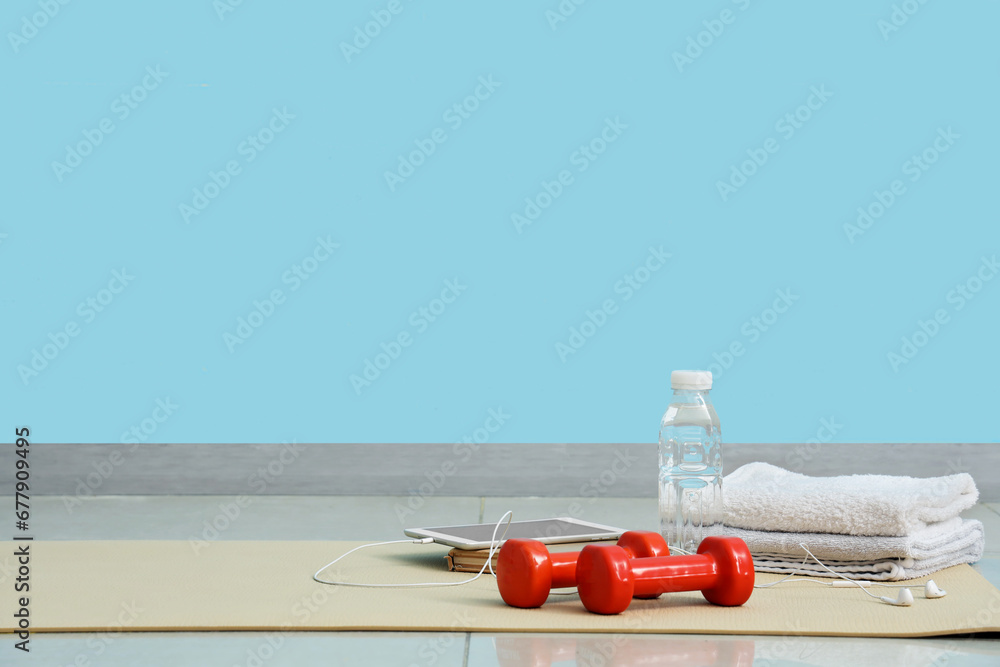 Tablet computer with dumbbells, bottle of water and towels on fitness mat in gym