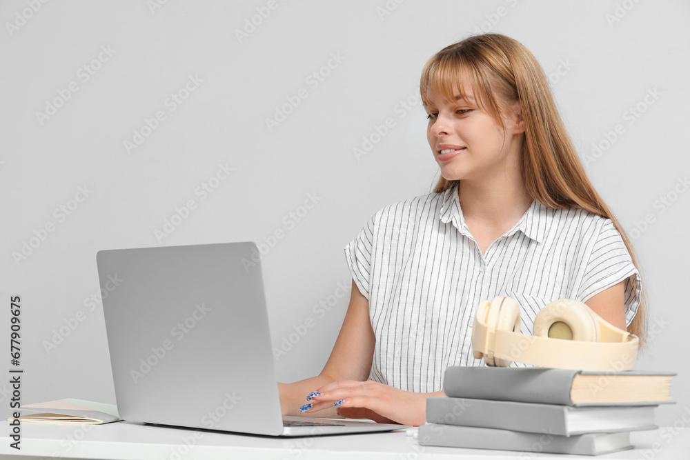 Female student with laptop doing homework at table on light background