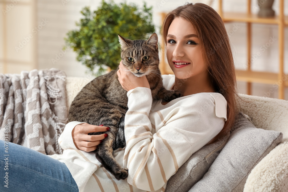 Pretty young woman holding cute tabby cat sitting on sofa at home