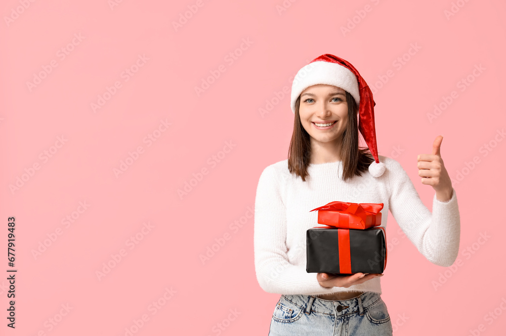 Beautiful young woman in Santa hat with gift boxes showing thumb-up on pink background