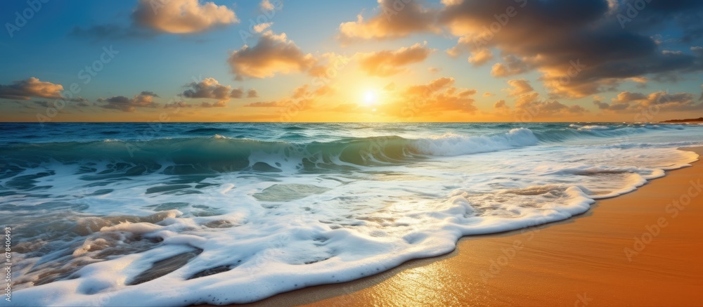 The breathtaking summer landscape of the beach with its azure blue sea golden sandy shores and vibrant orange hues of the setting sun against a backdrop of clear blue skies and fluffy white