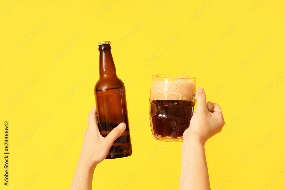 Female hands with bottle and mug of cold beer on yellow background