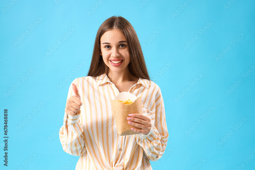 Beautiful happy young woman with pack of french fries showing thumb-up gesture on blue background