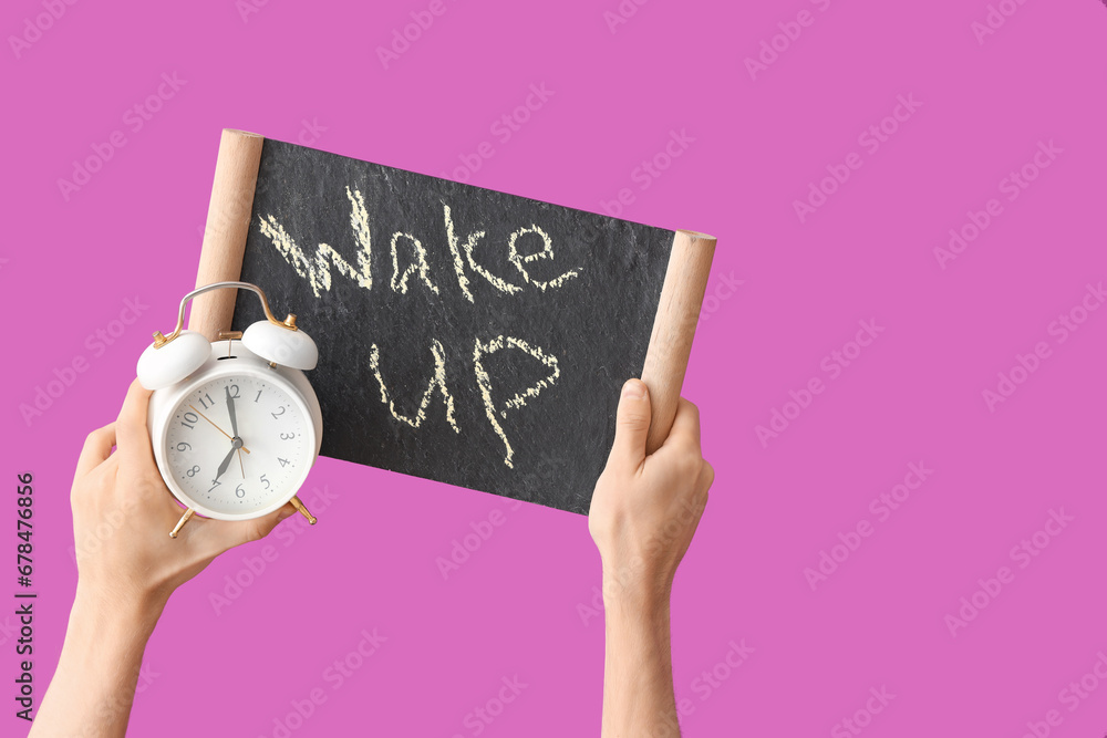 Male hands holding alarm clock and board with text WAKE UP on purple background