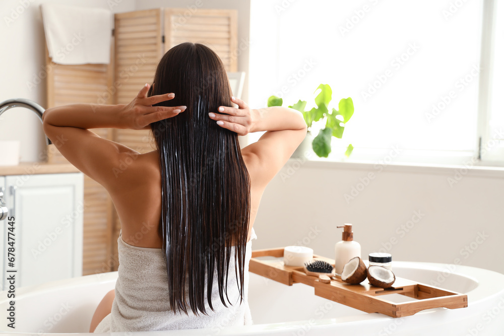 Beautiful young woman applying coconut oil onto her hair in bathroom, back view