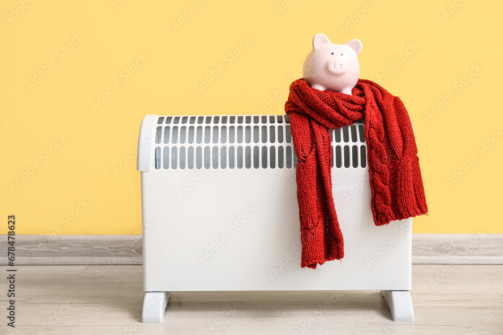 Piggy bank with warm scarf on electric convector heater near yellow wall at home. Heating saving concept