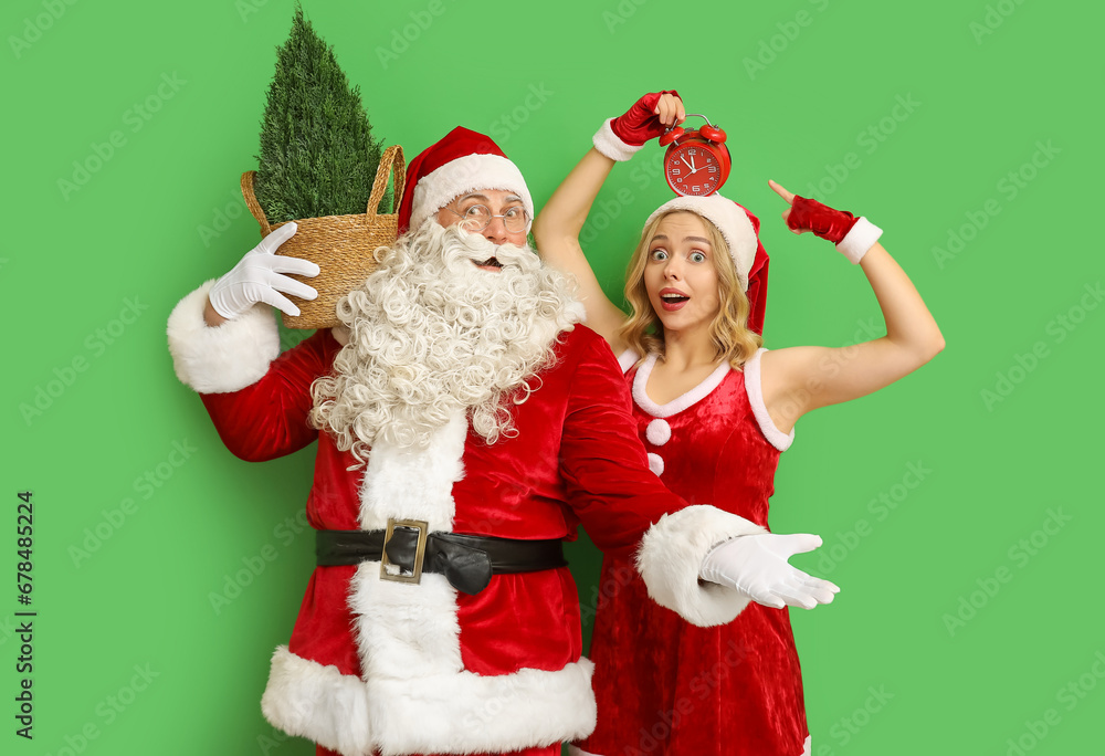 Surprised people dressed as Santa Claus with Cypress tree pointing at alarm clock on green background