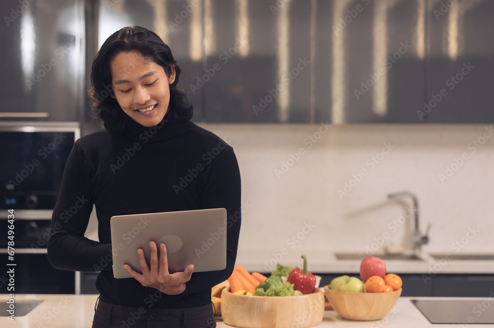 Asian man uses laptop computer and prepares healthy vegan salad in kitchen