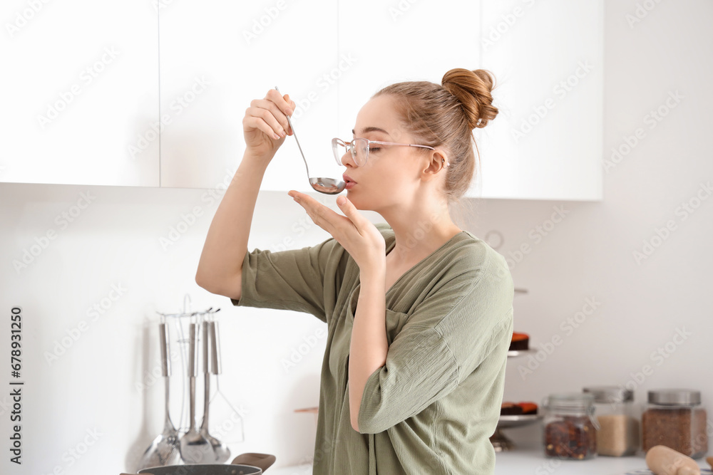 Pretty young woman tasting food from ladle in light kitchen