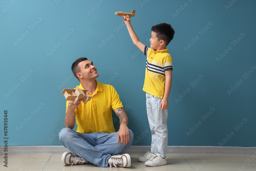 Young man and his little son playing with wooden airplanes near blue wall