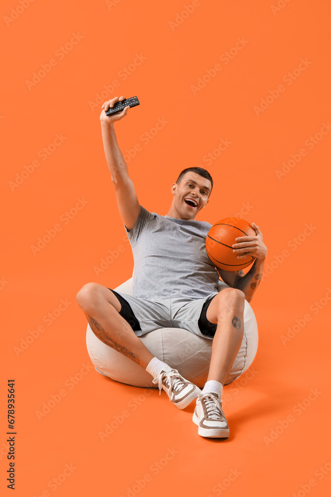 Young man with ball watching sport game on orange background