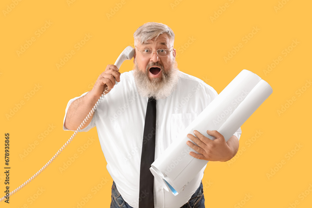 Mature shocked businessman with rolls of paper talking by mobile phone on yellow background