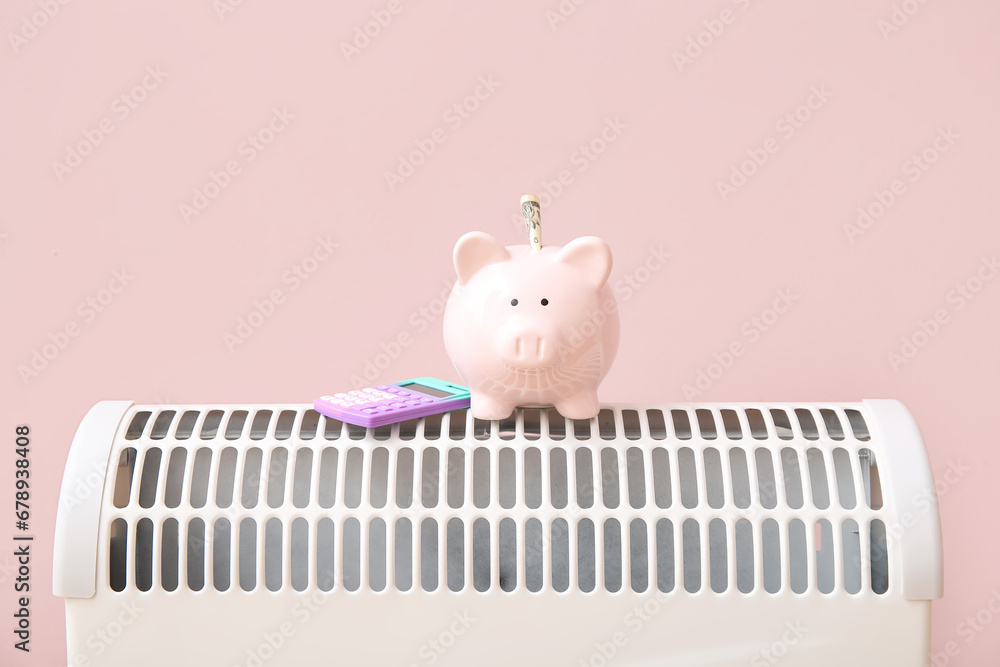 Piggy bank with calculator on electric convector heater near pink wall at home. Heating saving concept